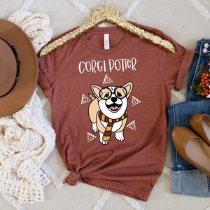 May include: A rust-colored t-shirt with a cartoon image of a corgi wearing glasses and a scarf. The text "Corgi Potter" is printed on the shirt.