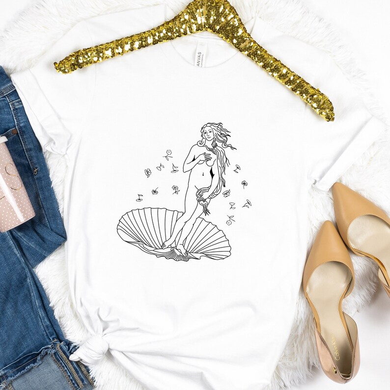 May include: A white t-shirt with a black line drawing of the Birth of Venus, a famous Renaissance painting. The image shows a woman standing on a seashell with her hair flowing behind her.