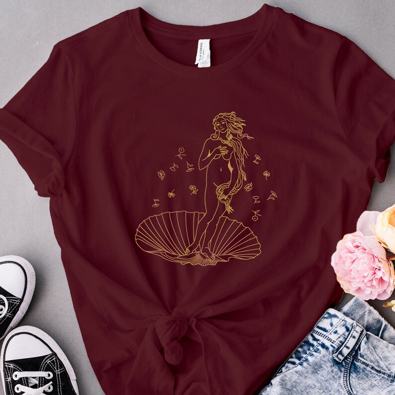 May include: A maroon t-shirt with a gold outline of the Birth of Venus, a famous Renaissance painting, printed on the front.