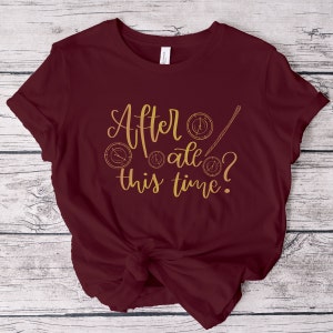 A picture showing a layed out Bella Canvas Unisex 3001 t-shirt in maroon color with the words after all this time; in cursive gold lettering and with wizard symbols.