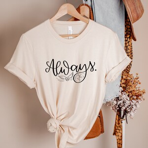 A picture showing a Bella Canvas Unisex 3001 t-shirt hanging on a rack in soft cream color with the word always; in cursive black lettering and with wizard symbols.