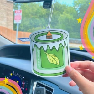 Può includere: Un profumatore per auto verde a forma di candela con un design a foglia. Il profumatore dice "Amber Standards" sul lato.