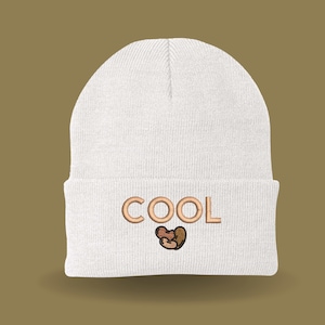 May include: White knit beanie with the word "COOL" embroidered in brown on the front. A small brown heart is below the word.