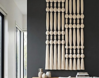 May include: A large wall hanging made of off-white fiber, featuring a series of vertical strands with alternating round knots and elongated cylindrical sections, ending in long, straight tassels suspended from a horizontal wooden rod.