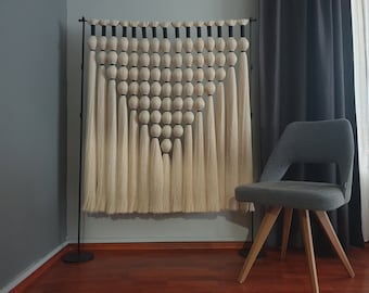May include: A large wall hanging composed of off-white yarn spheres and long, straight tassels suspended from a black metal rod. The spheres are arranged in a triangular pattern, with each section separated by black yarn wraps, creating a geometric design.