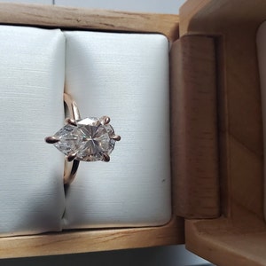 May include: A pear-shaped diamond engagement ring set in a rose gold band, displayed in a wooden box with the words "BRILLIANT EARTH" printed on the side.