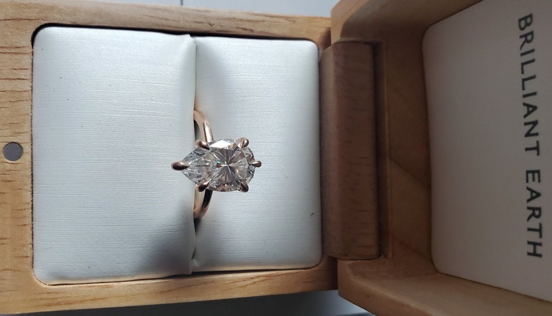 May include: A pear-shaped diamond engagement ring set in a rose gold band, displayed in a wooden box with the words "BRILLIANT EARTH" printed on the side.