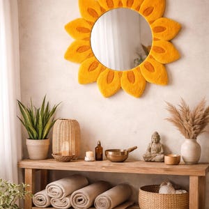 Op de afbeelding: Een decoratieve zonnebloemspiegel met gele vilten bloemblaadjes hangt boven een houten plank. De plank toont opgerolde handdoeken, planten, kaarsen en een Boeddha-beeld, wat een spa-achtige sfeer creëert.