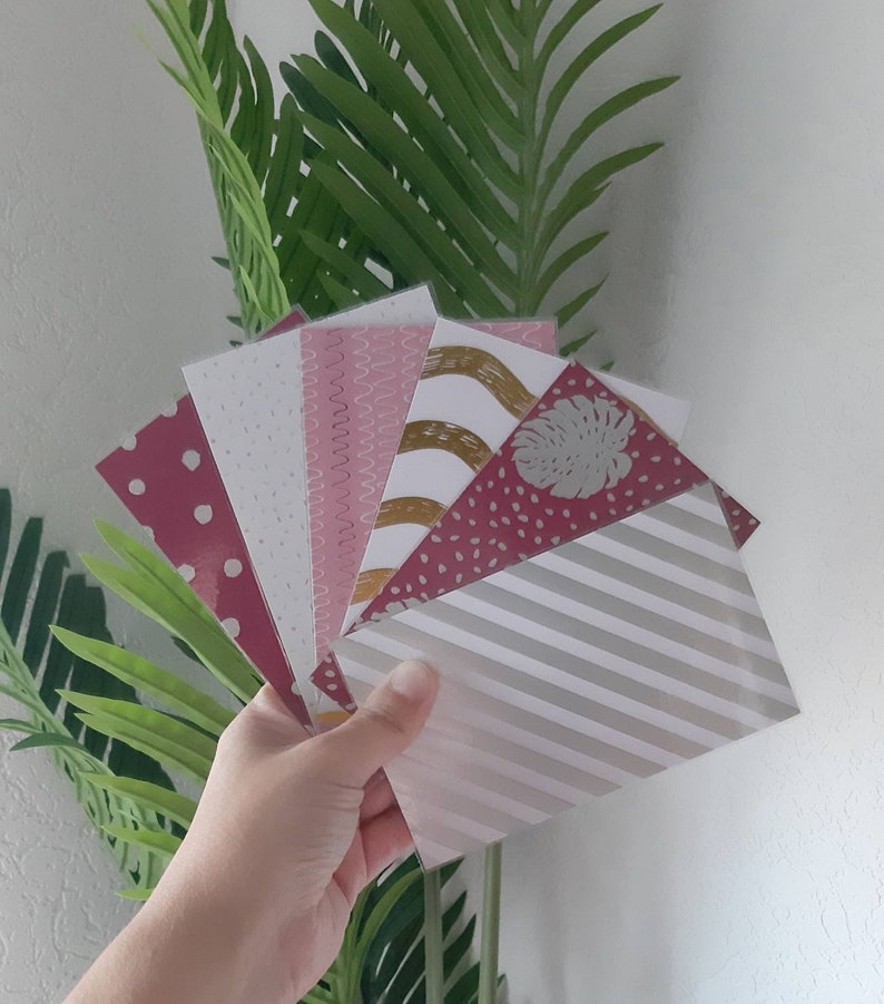 Puede incluir: Una mano sostiene un abanico de sobres de papel estampados. Los sobres presentan varios dise&ntilde;os, incluyendo lunares, l&iacute;neas onduladas y rayas, en tonos rosa, blanco y dorado. Se ve follaje verde en el fondo.