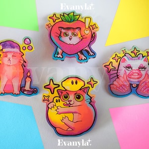 May include: Four colorful enamel pins featuring cartoon cats in various poses. The pins are on a white background with colorful paper behind them. The pins feature a cat wearing a witch's hat, a cat in a strawberry, a cat with a yellow halo, and a cat with pink manicured claws.