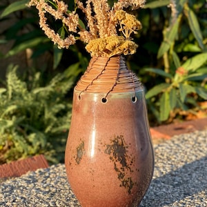 May include: A handcrafted ceramic vase with a reddish-brown glaze and dark brown accents. The vase has a unique shape and is adorned with a woven, light brown material around the neck. Dried flowers are arranged inside.