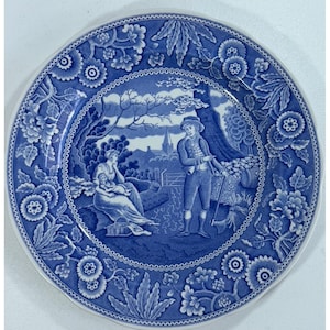 Puede incluir: Un plato redondo de cerámica con un patrón de transferencia azul y blanco. El centro representa una escena rural con una figura sentada sosteniendo a un niño, una figura de pie con un hacha y una iglesia a lo lejos. El borde tiene motivos florales.