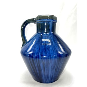 Può includere: Una brocca in ceramica con finitura lucida e una smaltatura a striature blu scuro e nere. Il recipiente presenta un corpo ampio e arrotondato che si restringe verso un collo stretto, con un manico curvo che parte dal lato fino al bordo.