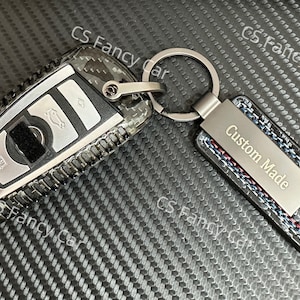 May include: A black and silver car key with a carbon fibre pattern and a silver key ring attached to a black leather keychain with the text "Custom Made" engraved on a silver plate.