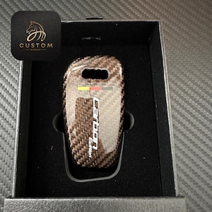 Può includere: Un portachiavi in fibra di carbonio nero con un logo "Carbon Edition" bianco e un design a strisce rosso, giallo e nero.