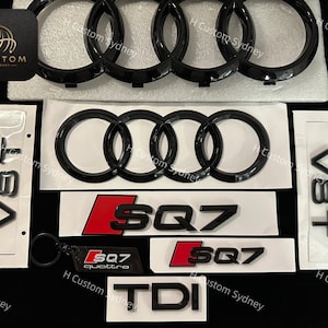 Könnte beinhalten: Ein Satz schwarzer und silberner Autoembleme. Die Embleme beinhalten die Audi-Ringe, "SQ7", "V8T" und "TDI".