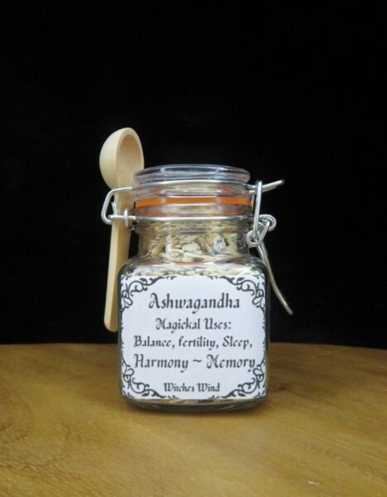 May include: A glass jar with a white label that reads "Ashwagandha Magickal Uses: Balance, fertility, Sleep, Harmony - Memory Witches Wind". The jar contains dried herbs and is accompanied by a wooden spoon.