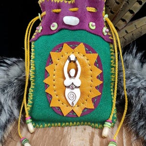 May include: A drawstring pouch made of green and purple felt, featuring a yellow leather sunburst applique with a white carved female figure at the center. The pouch includes yellow cords with beads, a white stone accent, and the text Witches Wind.