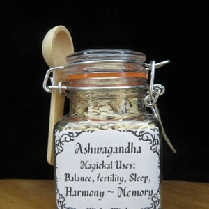 May include: A glass jar with a white label that reads "Ashwagandha Magickal Uses: Balance, fertility, Sleep, Harmony - Memory Witches Wind". The jar contains a light yellow powder and is accompanied by a wooden spoon.