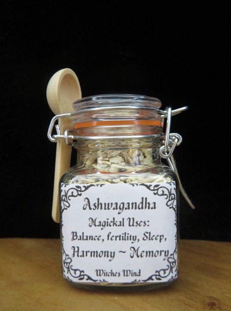 May include: A glass jar with a white label that reads "Ashwagandha Magickal Uses: Balance, fertility, Sleep, Harmony - Memory Witches Wind". The jar contains a light yellow powder and is accompanied by a wooden spoon.