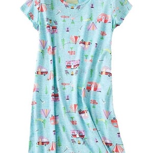 May include: A light blue, short-sleeved dress with a camping-themed pattern. The print includes red and pink camper vans, tents, trees, and camping chairs. The dress has a round neckline and a relaxed fit.