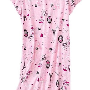 May include: A pink short-sleeved nightgown with a round neckline. The fabric is covered in a repeating pattern of Parisian-themed illustrations, including the Eiffel Tower, umbrellas, and portraits. The nightgown has a relaxed fit.
