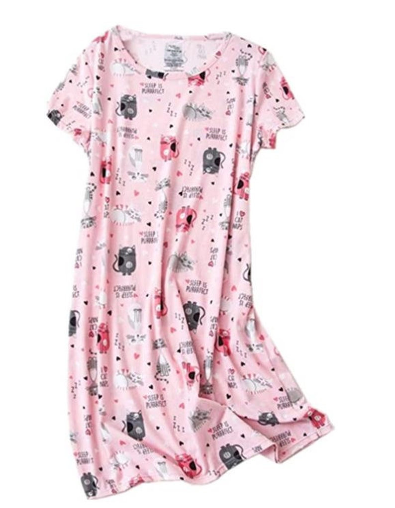 May include: A pink short-sleeved nightgown with a pattern of cartoon cats, hearts, and the phrase "Sleep is Purrfect". The cats are in various poses and colors, including gray, black, and red. The nightgown is made of a soft fabric.