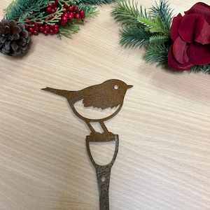 May include: A rusty metal silhouette of a robin perched on a post. The robin is facing right and has a white breast. The post has a rounded top and a long, thin stem.