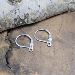 Könnte beinhalten: Zwei silberfarbene Ohrringverschlüsse werden auf einer Holzoberfläche gezeigt. Jeder Verschluss hat ein gebogenes, scharnierartiges Design mit einer kleinen Schlaufe an der Unterseite. Die Verschlüsse sind aus glänzendem Metall gefertigt und für die Schmuckherstellung bestimmt.