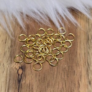 Könnte beinhalten: Eine Ansammlung kleiner, glänzender, goldfarbener Sprungringe auf einer Holzoberfläche. Die Ringe sind kreisförmig und scheinen aus Metall zu bestehen. Im Hintergrund befindet sich ein flauschiges, weißes Material.