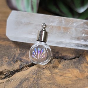Könnte beinhalten: Ein kleines, klares Glasfläschchen mit einem silberfarbenen Metallverschluss. Das Fläschchen hat ein irisierendes Design im Inneren. Das Fläschchen steht auf einer Holzoberfläche.
