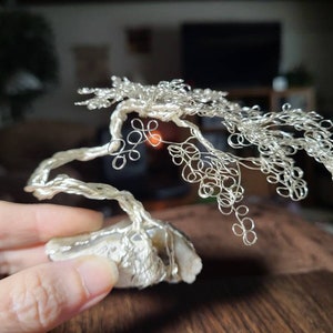 May include: A silver wire sculpture of a bonsai tree with a natural stone base. The tree is made of many small, intricate loops of wire, creating a delicate and realistic appearance.