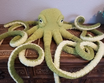 May include: A crocheted green octopus with white accents. The octopus has eight tentacles and is lying on a wooden surface.
