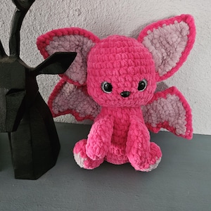 May include: A handmade pink crochet creature with large, light pink and gray wings. The plush toy has black eyes and a small black nose. The creature is sitting on a gray surface, with a black deer head sculpture in the background.