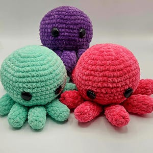 May include: Three plush octopuses in shades of purple, teal, and pink. Each has a round body, eight tentacles, and large black button eyes. The soft, textured material gives them a cuddly appearance.
