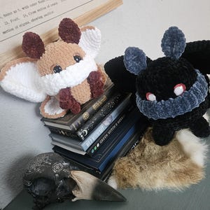 May include: Two plush moth toys made of soft velvet yarn. One moth is tan and white with dark brown accents, while the other is black with a dark gray neck ruff and red eyes. They are positioned on a stack of books next to a dark bird skull.