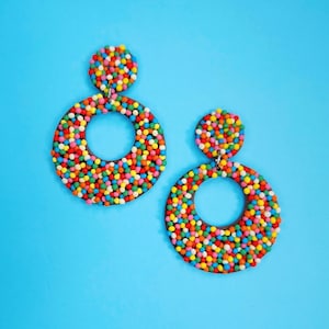 May include: A pair of dangle earrings featuring a circular design covered in tiny, multicolored spherical beads. Each earring consists of a small solid circle attached to a larger open hoop, both densely textured with vibrant red, yellow, blue, green, and orange dots.
