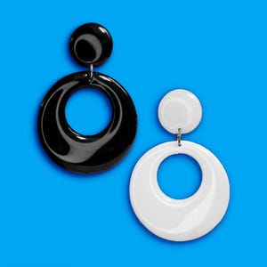 May include: A pair of retro-style earrings. One earring is black, and the other is white. Each earring features a small, round stud connected to a large, circular pendant. The earrings are set against a bright blue background.