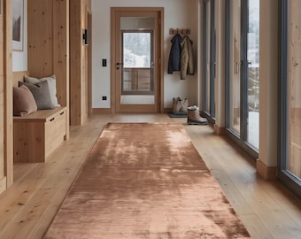 May include: A rectangular runner rug with a metallic copper-toned finish and a subtle, textured sheen. The rug is positioned centrally on a light-colored wooden floor within a hallway featuring light wood paneling and large glass windows.