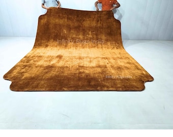 Puede incluir: Una alfombra rectangular grande de color marrón dorado intenso y sólido con una textura suave y afelpada. La alfombra presenta una forma distintiva e irregular con esquinas redondeadas y recortes en el borde inferior. El texto Infinity Rugs India es visible.
