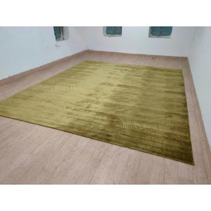 Puede incluir: Una alfombra grande, rectangular, de color verde oliva con un brillo sutil. La alfombra está colocada sobre un suelo de madera en una habitación con paredes blancas.