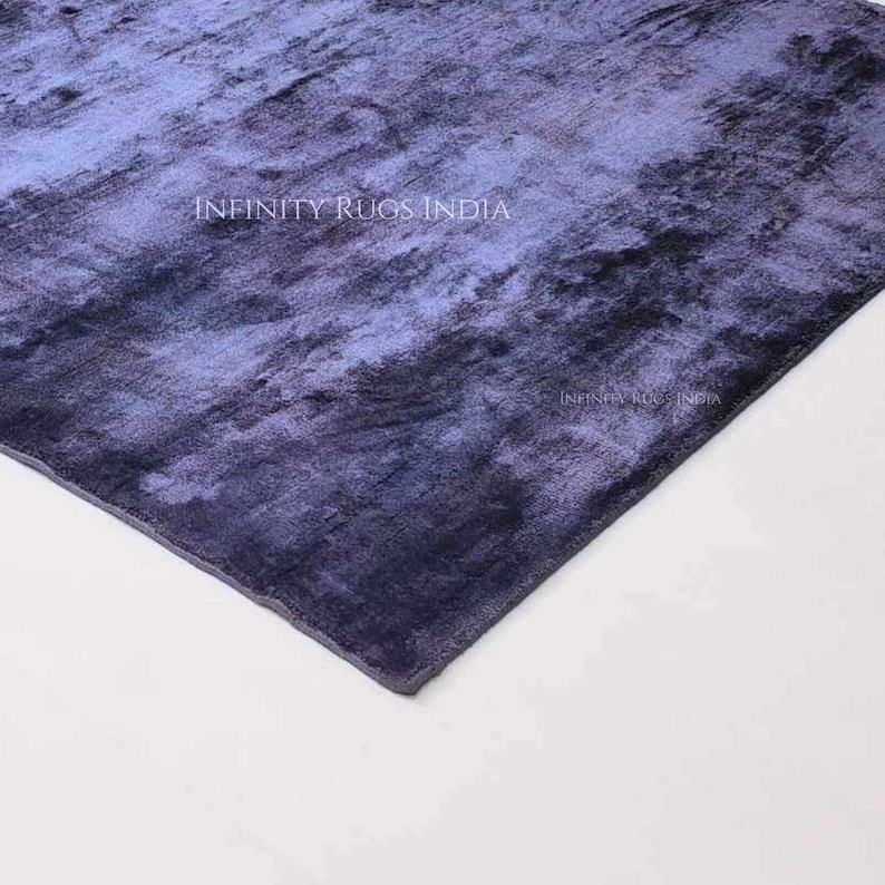 May include: A close-up of a blue and purple rug with a textured surface. The rug has a faded, abstract design. The text "INFINITY RUGS INDIA" is visible in the upper center of the rug.