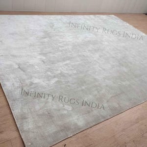 Puede incluir: Una alfombra grande de color gris claro con un patr&oacute;n sutil y texturizado. La alfombra est&aacute; colocada sobre un suelo de madera. El texto "INFINITY RUGS INDIA" est&aacute; impreso dos veces en la alfombra.