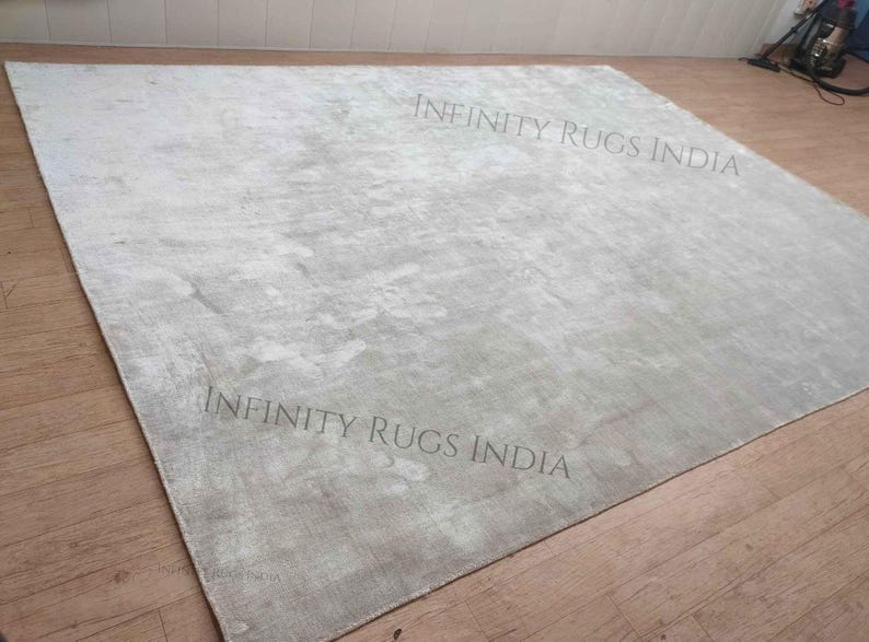 Puede incluir: Una alfombra grande de color gris claro con un patr&oacute;n sutil y texturizado. La alfombra est&aacute; colocada sobre un suelo de madera. El texto "INFINITY RUGS INDIA" est&aacute; impreso dos veces en la alfombra.