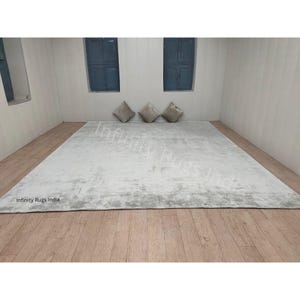 Puede incluir: Una alfombra rectangular blanca con un patrón sutil y desgastado. La alfombra está colocada sobre un suelo de madera en una habitación con dos ventanas y tres cojines decorativos.