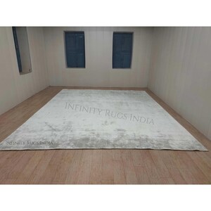 Puede incluir: Una alfombra de &aacute;rea blanca y gris con un sutil patr&oacute;n desgastado. La alfombra est&aacute; colocada sobre un suelo de madera en una habitaci&oacute;n con dos ventanas. El texto "INFINITY RUGS INDIA" es visible en la imagen.