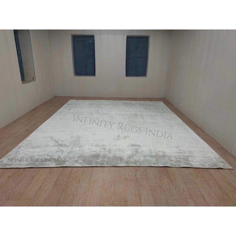 Puede incluir: Una alfombra de &aacute;rea blanca y gris con un sutil patr&oacute;n desgastado. La alfombra est&aacute; colocada sobre un suelo de madera en una habitaci&oacute;n con dos ventanas. El texto "INFINITY RUGS INDIA" es visible en la imagen.