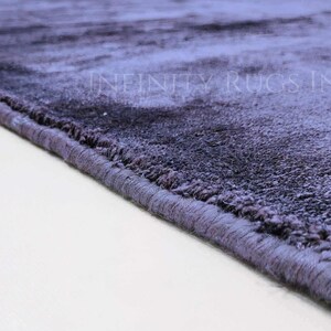May include: Close-up of the edge of a soft, plush, purple rug with a subtle ombre effect. The rug has a stitched edge and is made of a high-quality material.