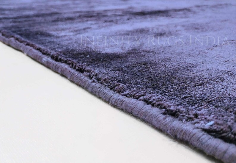May include: Close-up of the edge of a soft, plush, purple rug with a subtle ombre effect. The rug has a stitched edge and is made of a high-quality material.