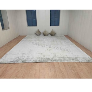 Puede incluir: Una alfombra rectangular blanca con un patr&oacute;n sutil y desgastado. La alfombra est&aacute; colocada sobre un suelo de madera en una habitaci&oacute;n con dos ventanas y tres almohadas. El texto "INFINITY RUGS INDIA" est&aacute; impreso en la alfombra.